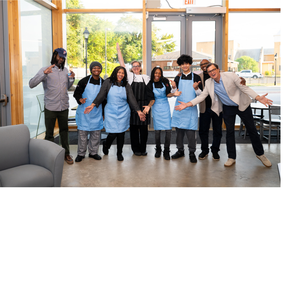 Group of diverse people wearing blue aprons pose together inside a bright storefront, smiling with arms around each other.