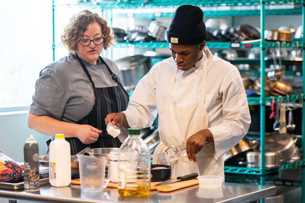 Two chefs in a stainless-steel kitchen preparing ingredients at a prep station with bottles and bowls nearby.