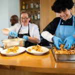 Two cooks in gloves serving shrimp from trays onto plates at a buffet counter, smiling as they work