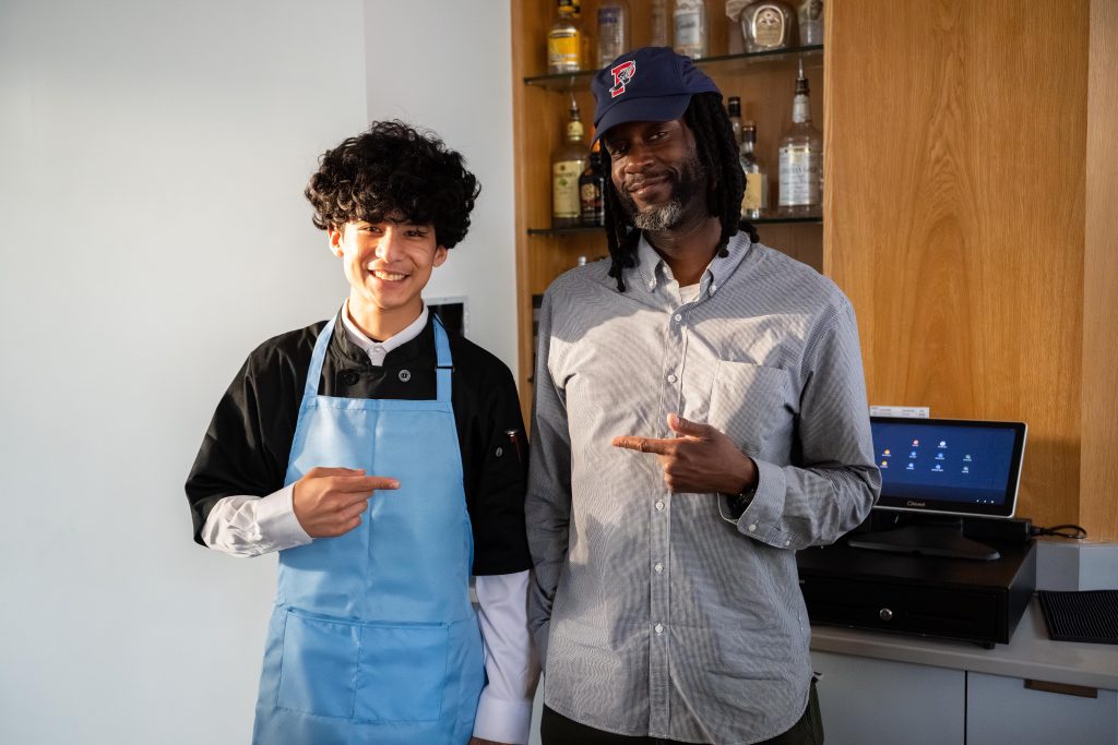 Two smiling men pose for the camera in a bar area, one in a blue apron and the other wearing a cap and checkered shirt, both pointing at each other.