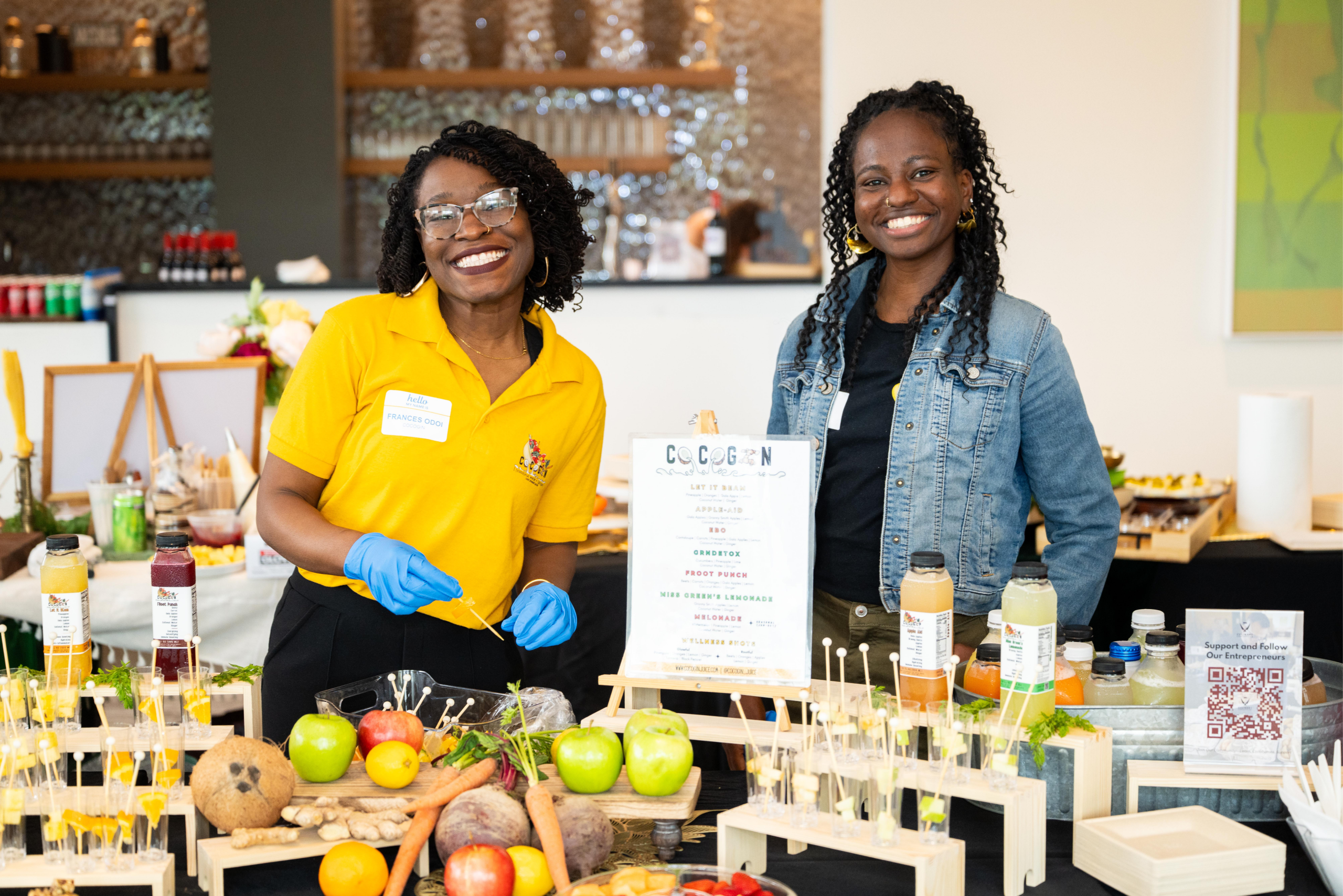 Untitled design (55) Two smiling women stand behind a colorful fruit and juice booth, with bottles, skewers, and fresh produce laid out on the counter in front of them.