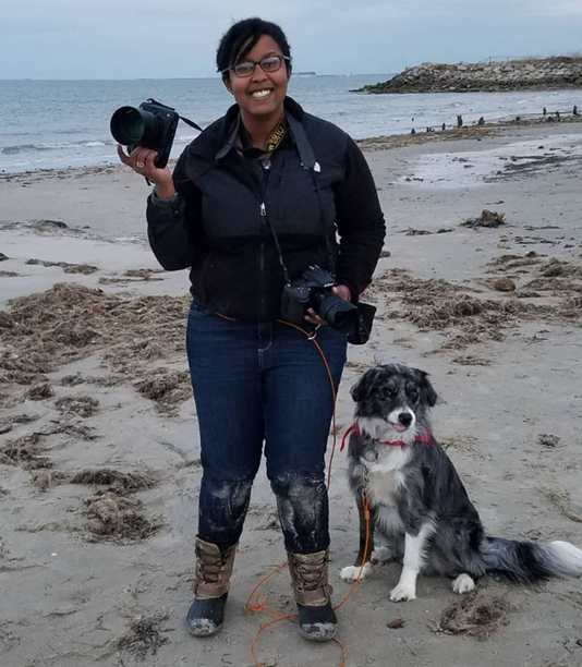 Photographer holding two cameras standing on a beach with her dog
